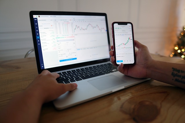 person's hands on a laptop displaying stocks, also holding a phone with sotcks on it